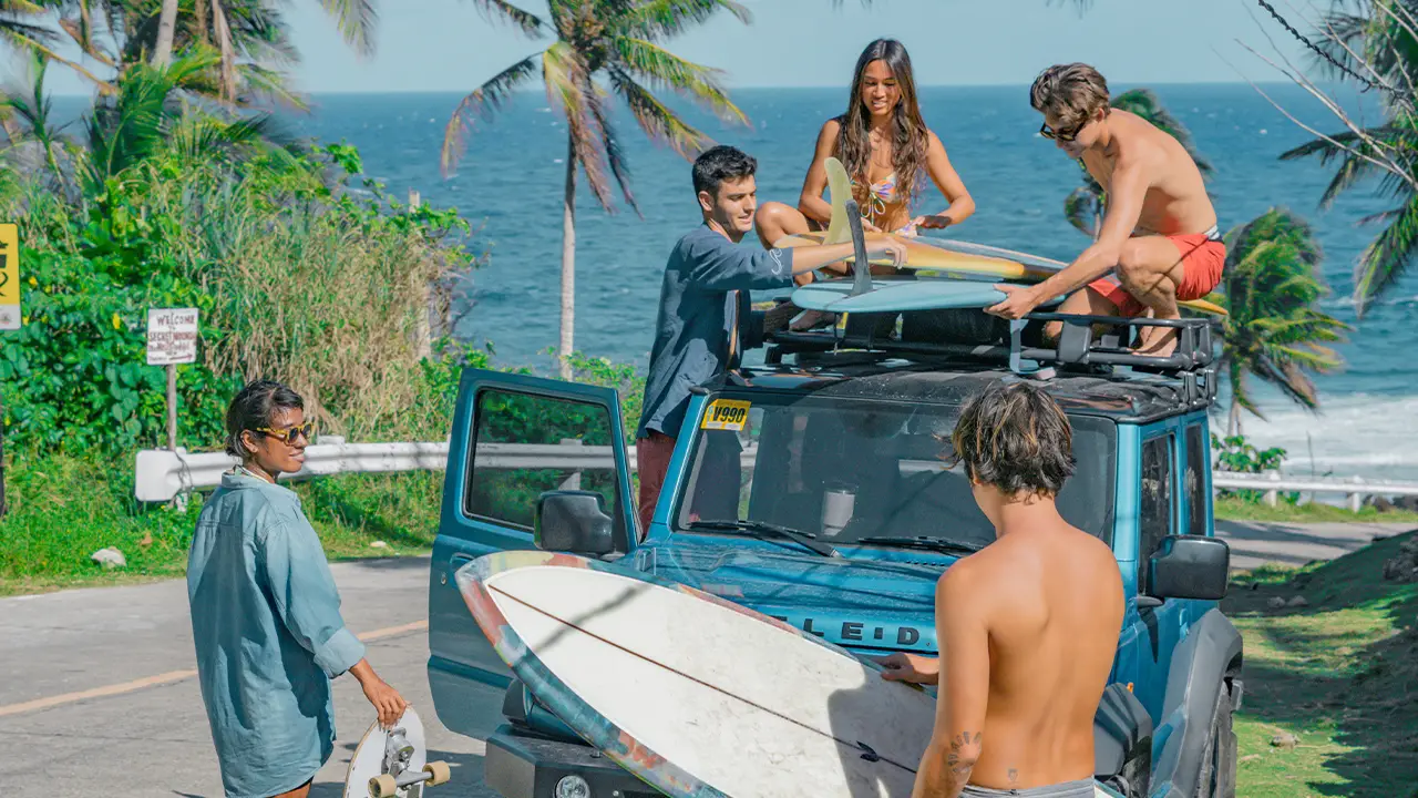 Jeep is parked on the hill while they're setting up their surfboards in top of their jeep with blue shining sea on their back