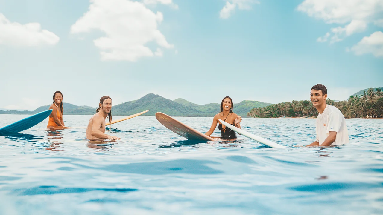 People having on the beautiful blue waters of siargao while on their surf boards, with a greenery coconuts and mountains in the background